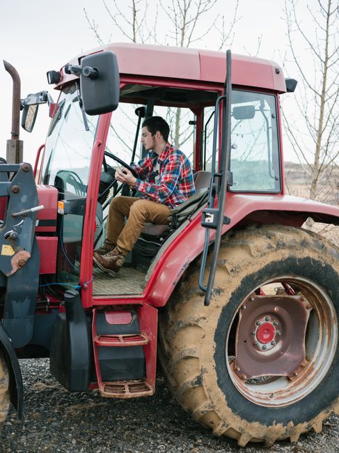 Adult man in checkered shirt driving tractor on a farm. Outdoors, daytime.